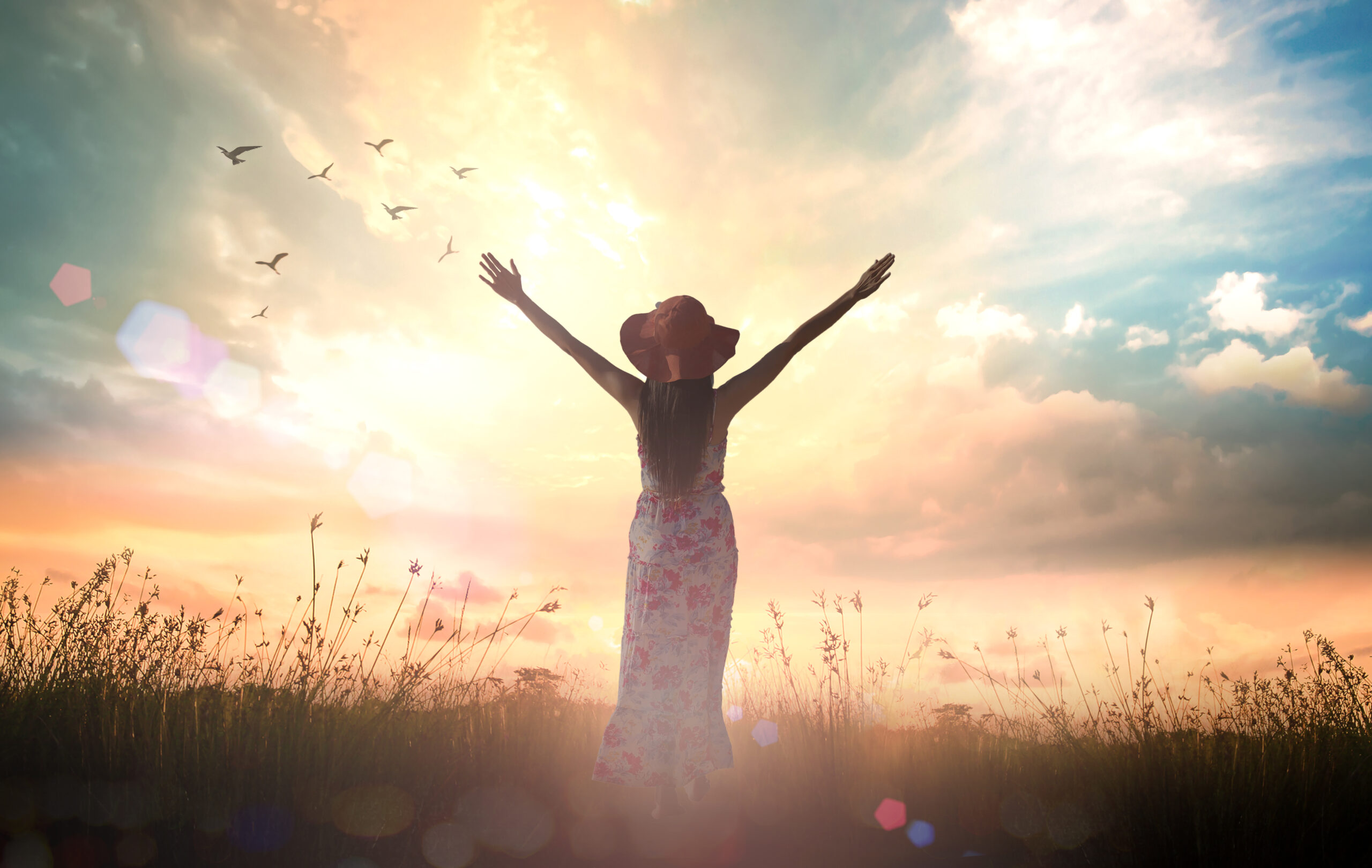 A woman facing a vibrant sunset with her arms outstretched, symbolizing freedom, abundance, and manifesting her dream life.