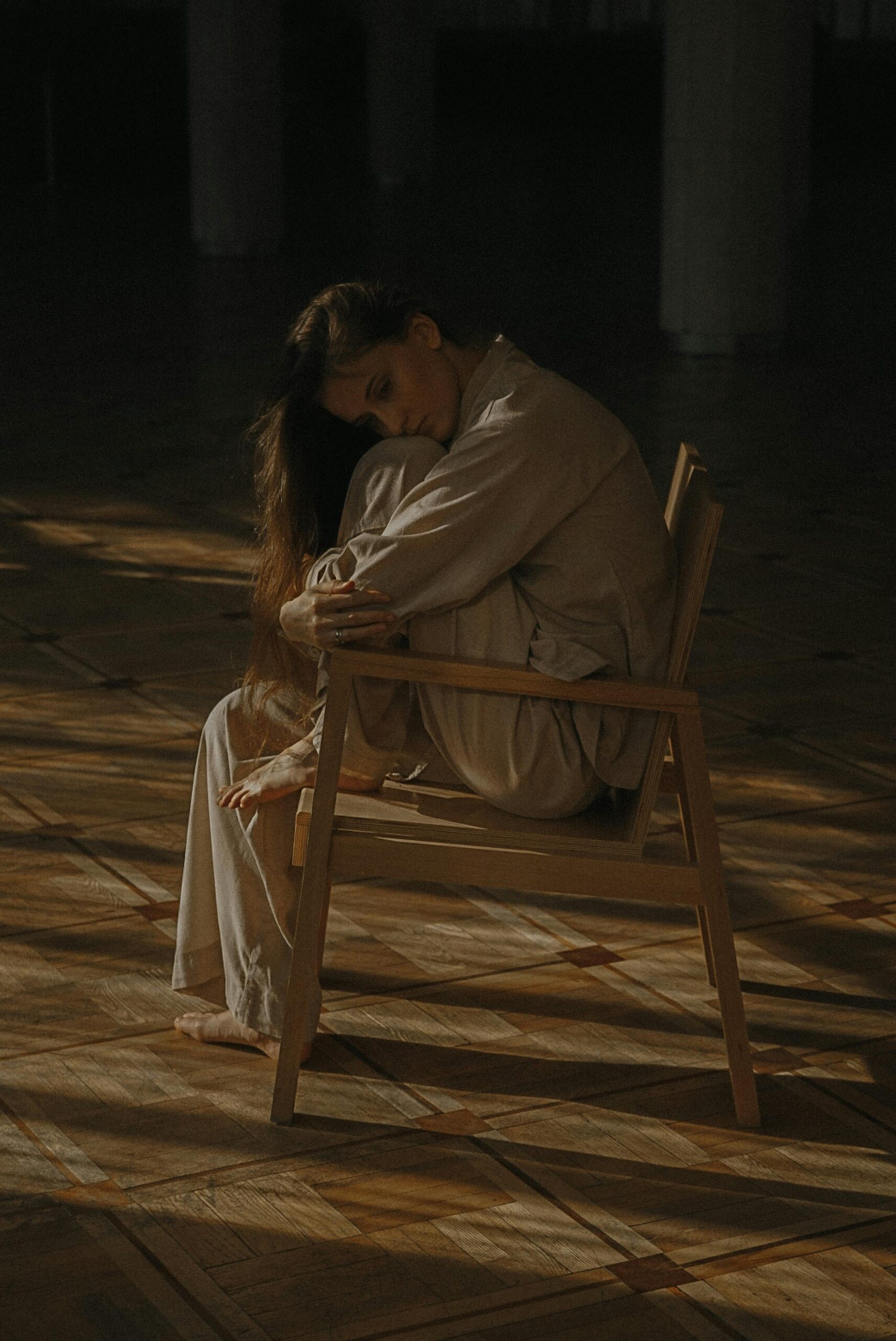 A woman sitting in a dimly lit room, looking sad and dissociating, representing the struggle of overcoming limiting beliefs to manifest your dream life.