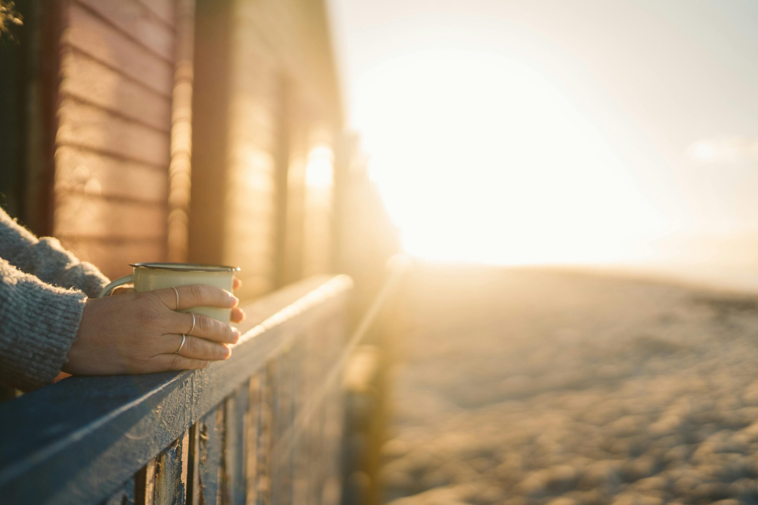 A woman standing on her porch with a cup of coffee in her hands, watching a peaceful sunrise, symbolizing the calm and clarity of how to start living intentionally.