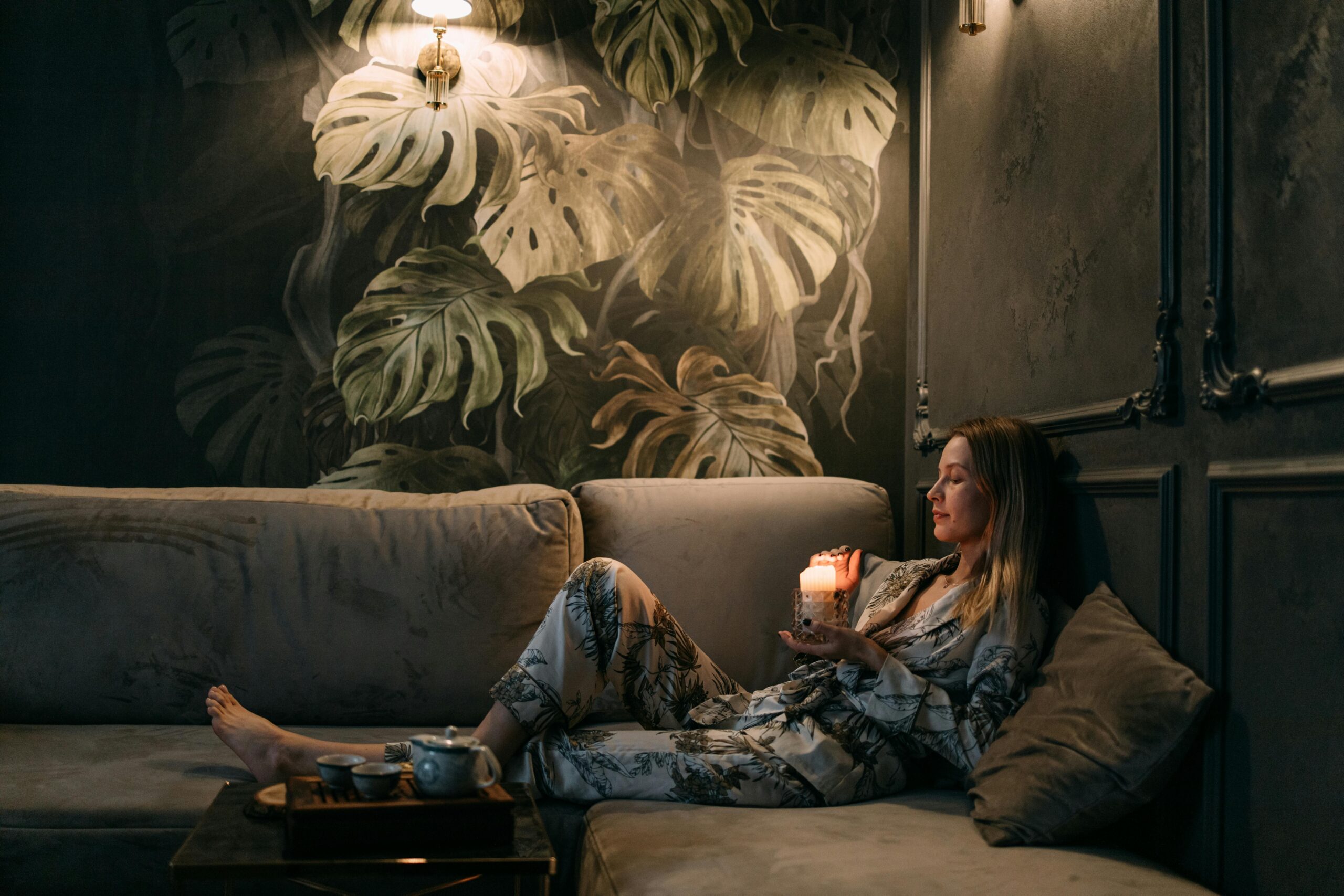 A woman in matching pajamas sits on a couch in her living room during the evening, holding a candle and feeling its warmth, with eyes closed as she practices breathwork.