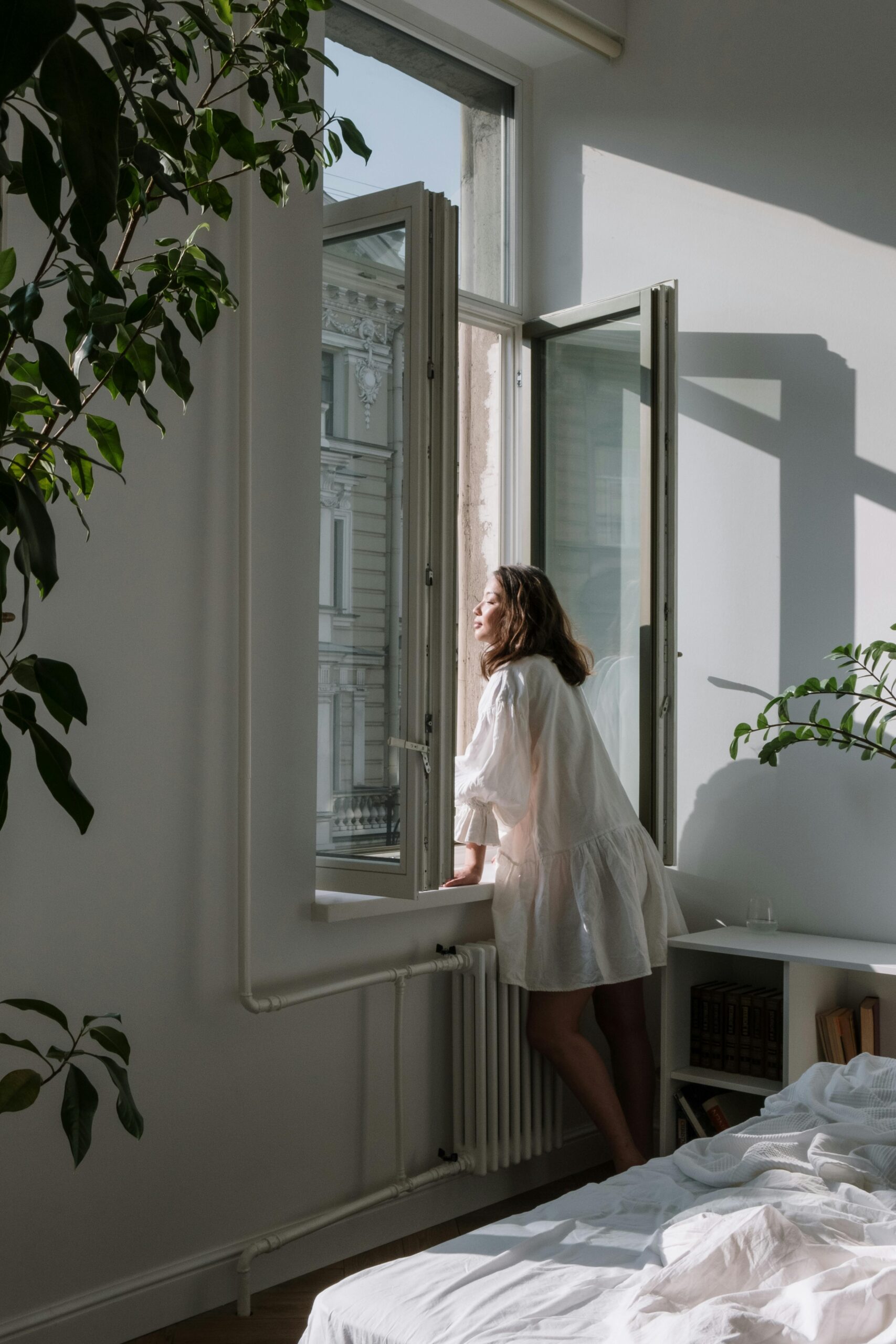 A woman standing in her bedroom, leaning slightly out of wide-open windows as she breathes in fresh air, symbolizing clarity and connection with breathwork for manifestation.