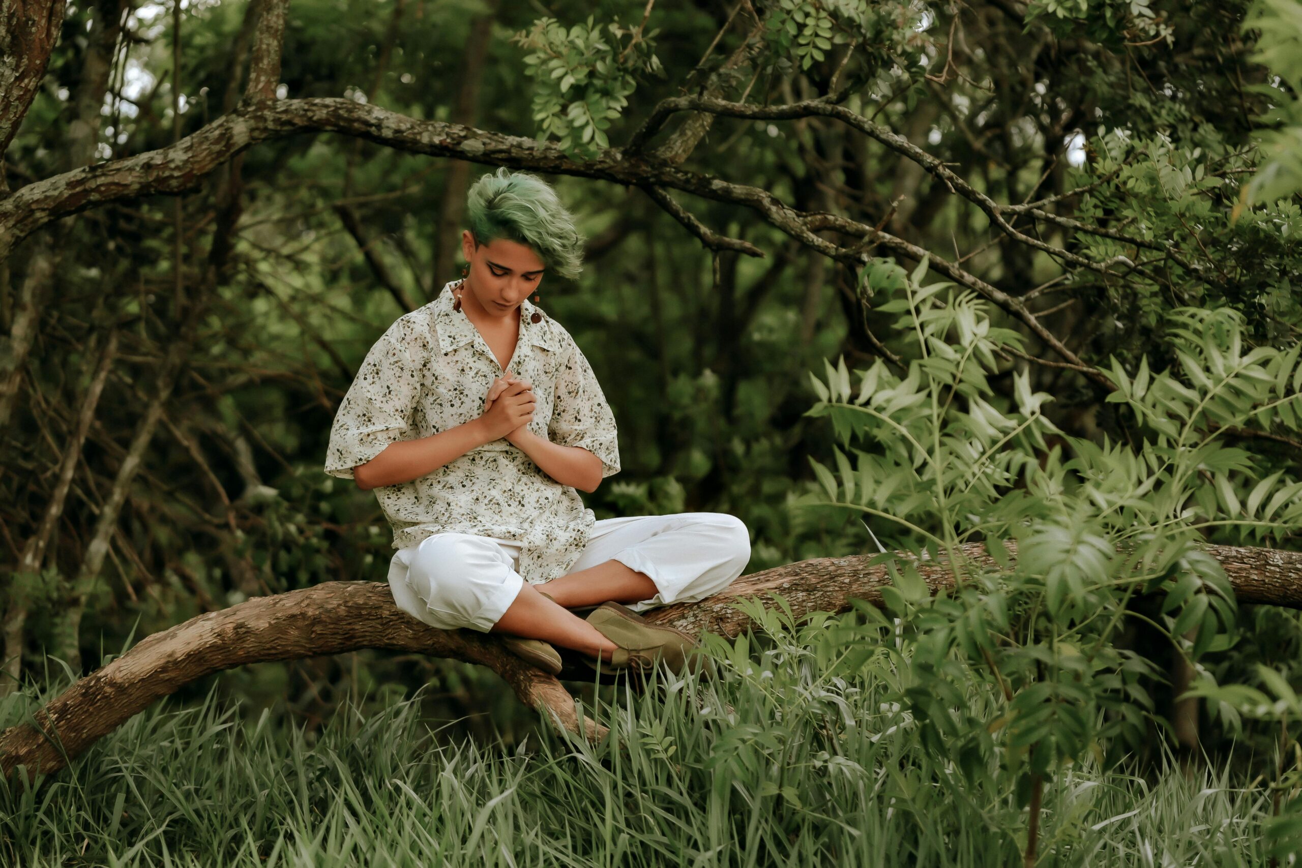A person sitting cross-legged on a low-hanging branch, surrounded by lush greenery, with hands resting on their heart while breathing deeply, symbolizing grounding and connection.