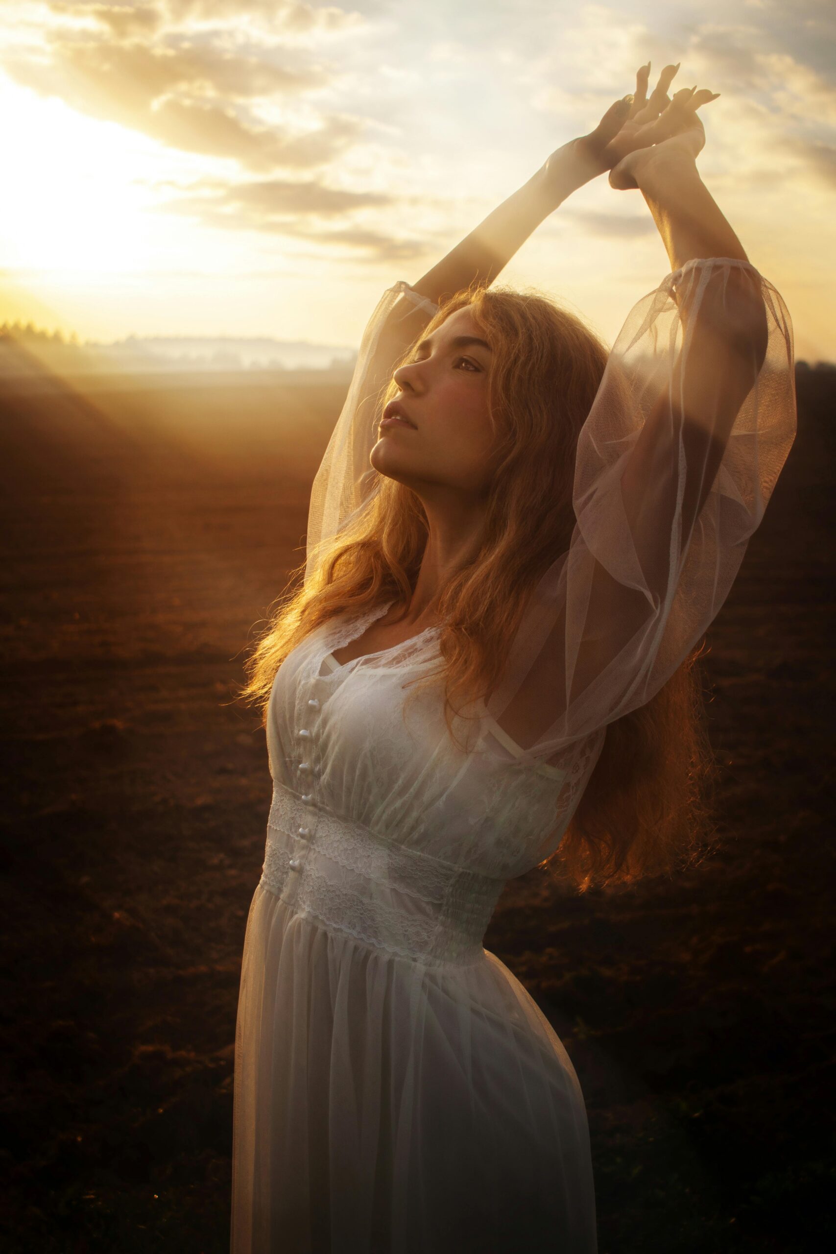 Feminine Energy Traits embodied by a woman in a flowy dress, standing in a field at golden hour, with arms raised above her head.