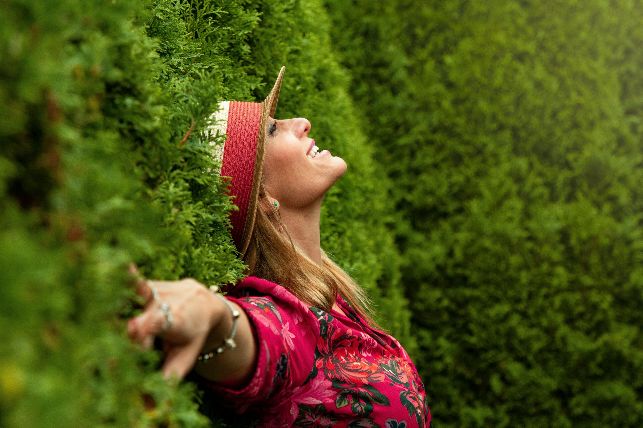 A woman standing outside, leaning against a lush green wall of bushes with her arms outstretched and a joyful smile toward the sky.