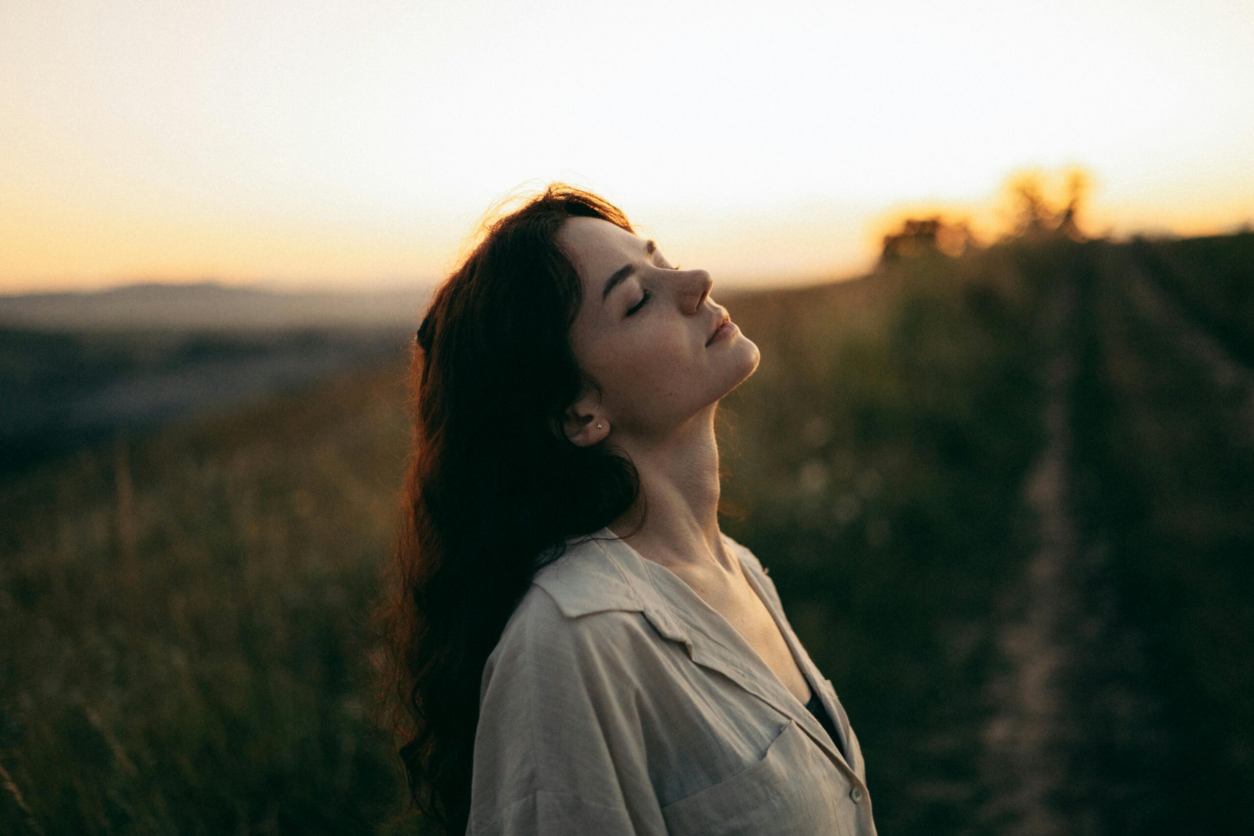 Serene woman in a field with eyes closed, head tilted towards the setting sun, symbolizing the Law of Detachment and inner peace.