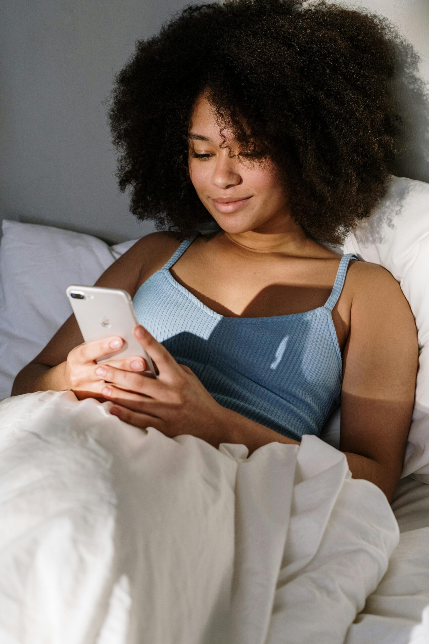 Young woman sitting in bed smiling while looking at her phone, representing joy and positive energy while learning how to manifest a text from someone.