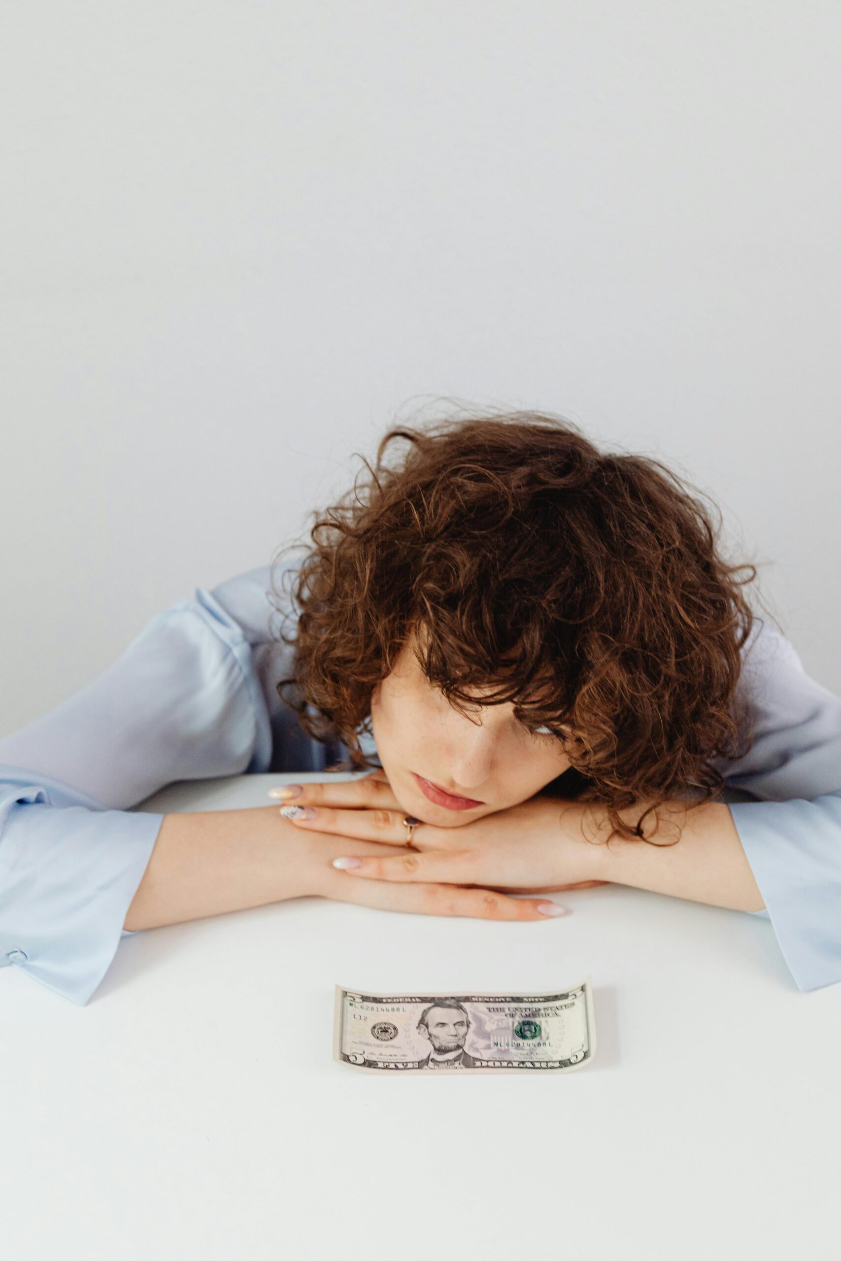 A worried woman rests her head on her hands, staring at a single $5 bill on the table, symbolizing financial stress and the need to release spiritual money blocks.