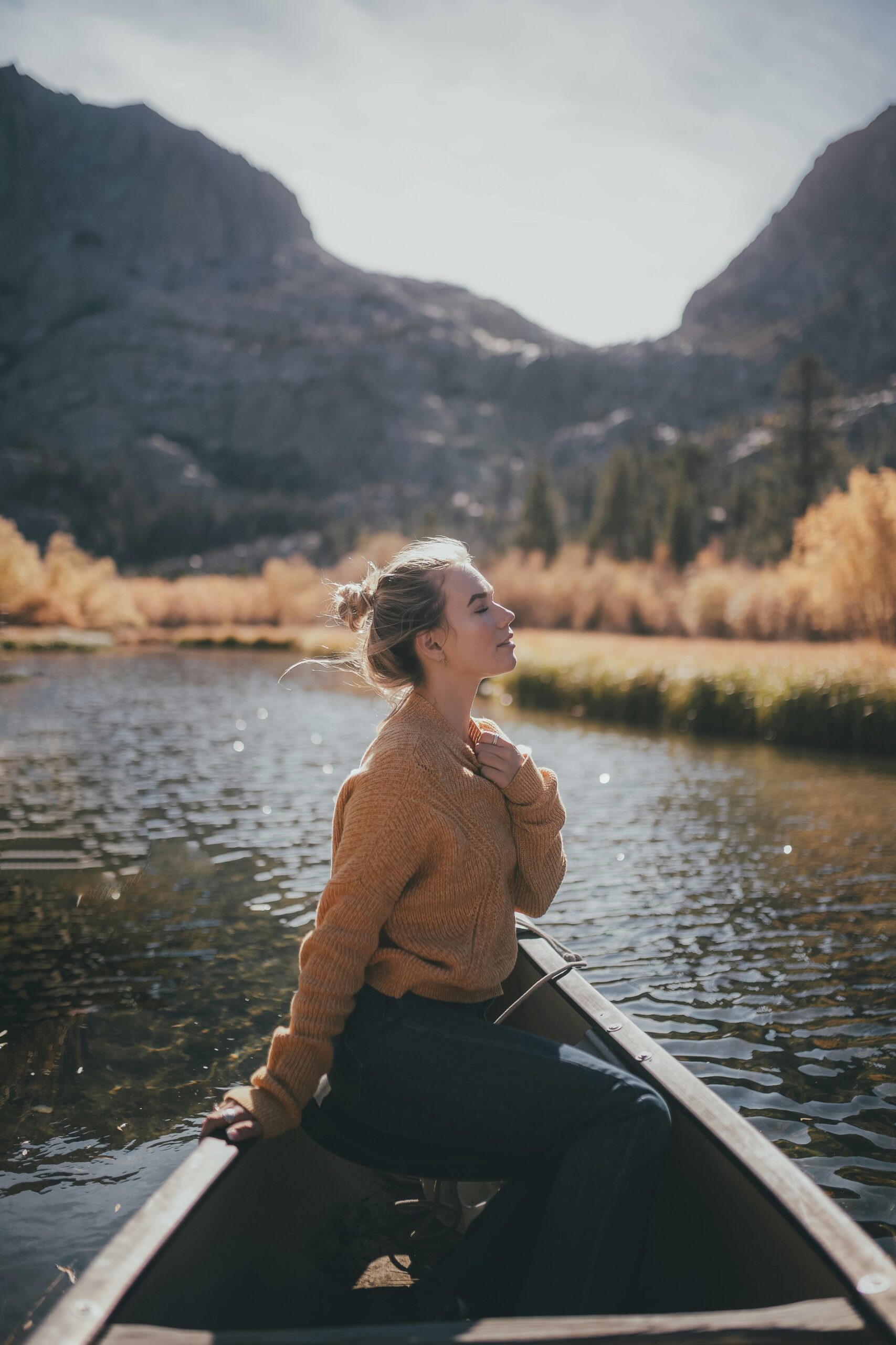 A woman sitting in a canoe on a peaceful river, hand on her heart, face lifted to the sun, symbolizing alignment and syncing with the universe.