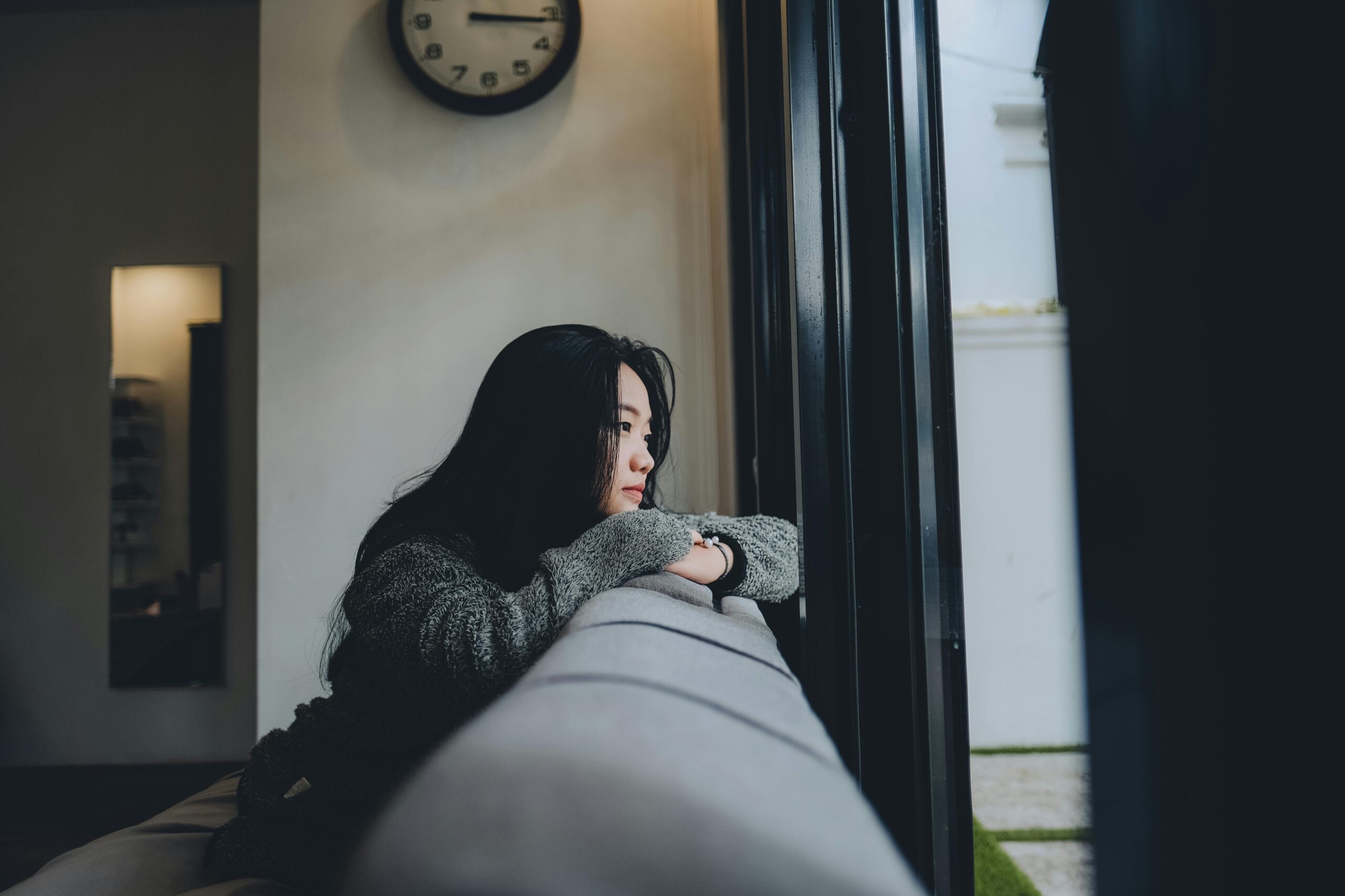 A woman leaning over her couch, looking out the window with a thoughtful expression, symbolizing waiting and trusting divine timing.