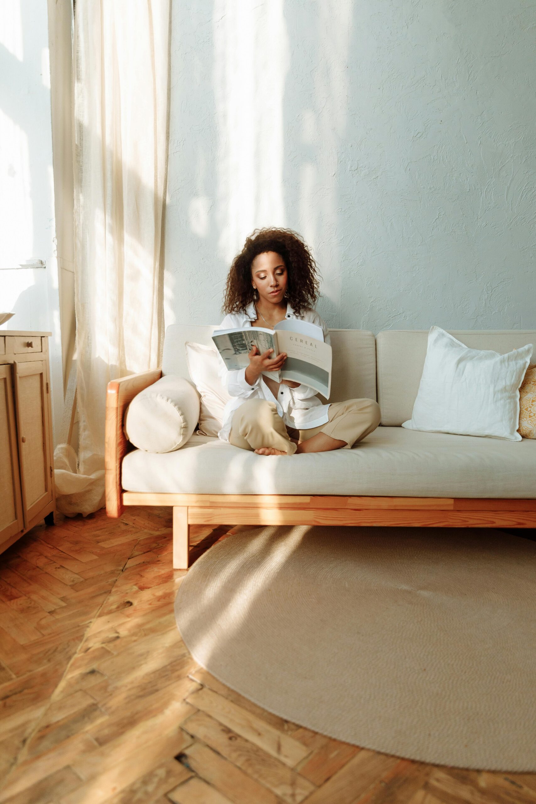 A minimalist living room with a cozy ambiance, featuring a woman sitting on her couch reading peacefully. This serene setting reflects the essence of slow living and intentionality. How to Live a More Simple Life starts with creating a calming space that nurtures peace and mindfulness.