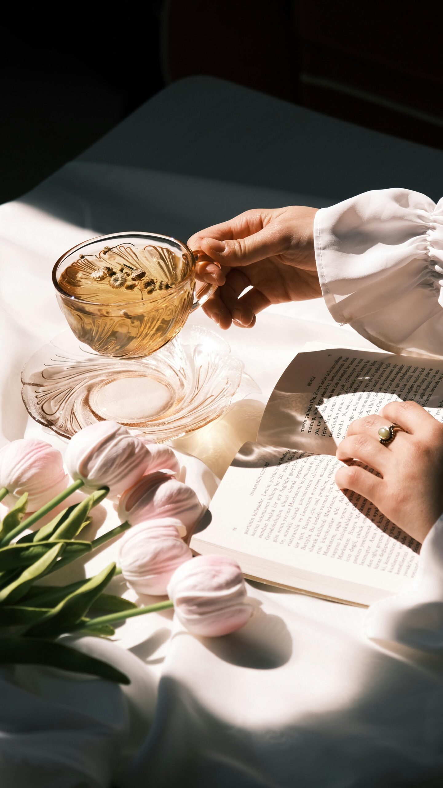 A woman practicing wealth manifestation by enjoying a cup of tea in the morning, reading next to beautiful flowers.