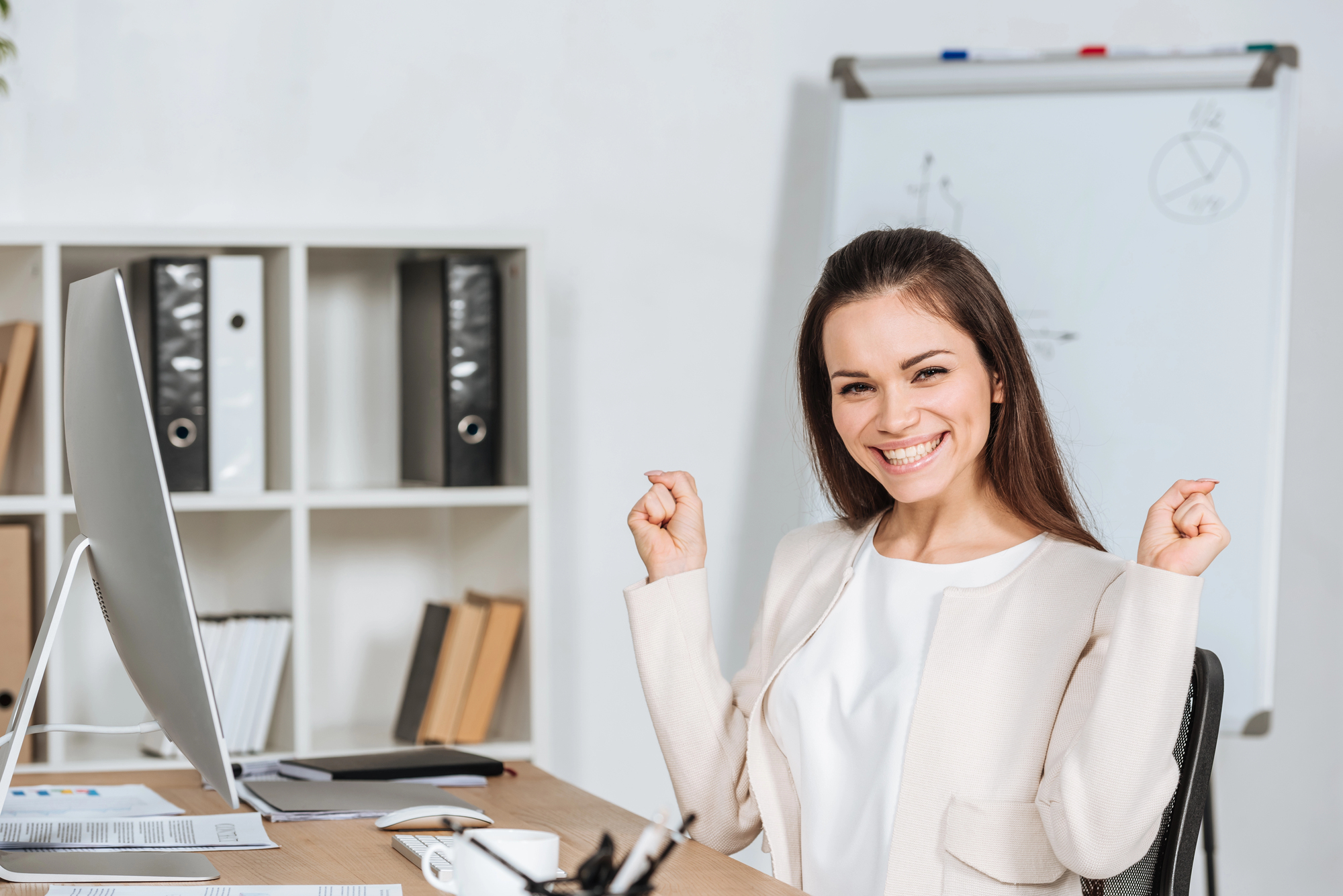 A joyful woman sitting at her desk with arms raised in victory, smiling brightly as she embodies the law of assumption and celebrates her desires manifesting into reality.